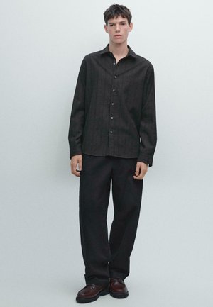 Giovane uomo in piedi con una camicia button-up scura, pantaloni neri larghi e scarpe di pelle marrone su uno sfondo chiaro e semplice.