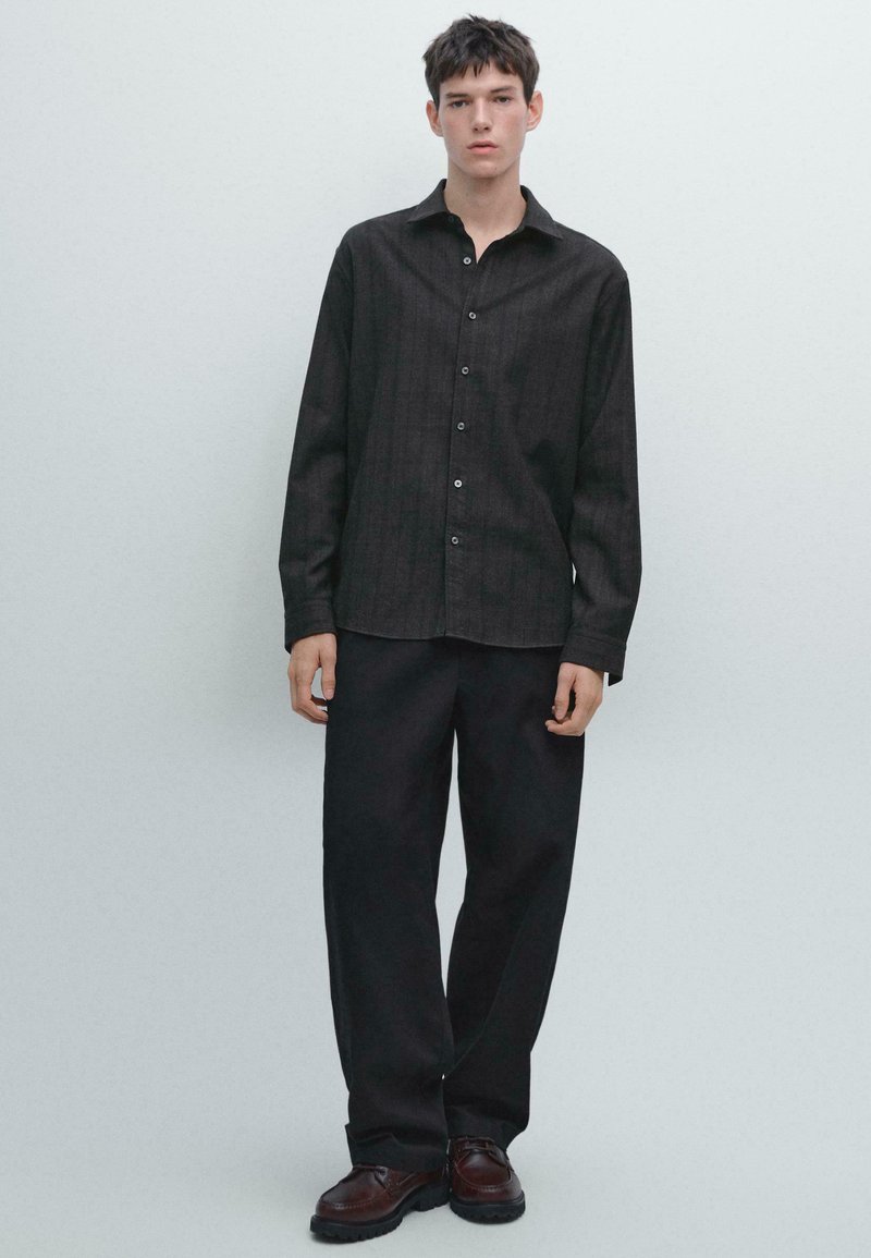 Ung mand står iført en mørk skjorte med knapper, løse sorte bukser og brune lædersko mod en ensfarvet lys baggrund.