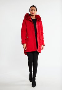 Parka rossa con cappuccio in pelliccia sintetica, due tasche frontali e chiusura con zip. Indossata sopra leggings neri e stivaletti.