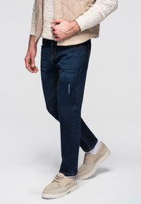 Jean en denim bleu foncé avec une coupe ajustée, légèrement usé, et des surpiqûres jaunes, associé à des chaussures beige et un gilet tricoté crème.