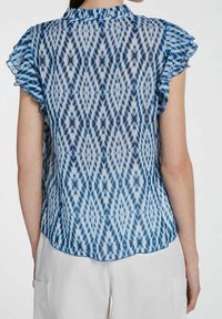 Blouse bleue à motifs avec manches volantées, en tissu léger et transparent, à ourlet arrondi. Détails complexes de motifs en diamant et floraux.