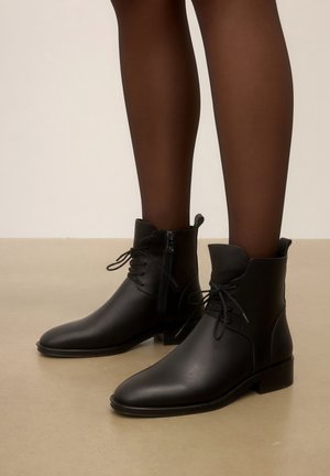 Pieds portant des bottines en cuir noir avec fermetures éclair latérales et lacets, associées à des collants noirs transparents sur un sol beige.