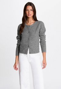 Frau in einer grauen, knöpfbaren Strickjacke mit langen Ärmeln und weißen Hosen mit kleinen Strassdetails am Bund.