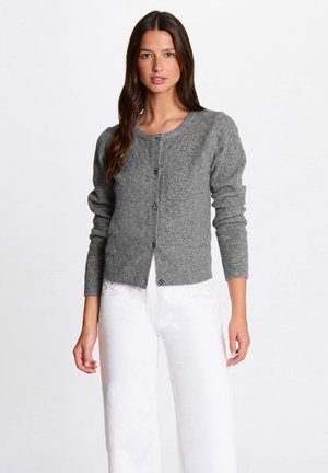 Frau in einer grauen, knöpfbaren Strickjacke mit langen Ärmeln und weißen Hosen mit kleinen Strassdetails am Bund.