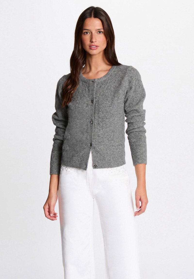 Frau in einer grauen, knöpfbaren Strickjacke mit langen Ärmeln und weißen Hosen mit kleinen Strassdetails am Bund.