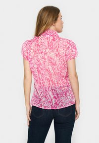 Short-sleeve blouse in varying shades of pink with a textured fabric, featuring a ruffled collar and abstract pattern. Fitted design at the waist.