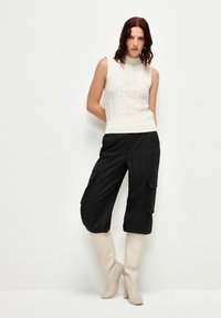 Maglione senza maniche in crema con motivi a trecce, abbinato a pantaloni cargo neri con righe verticali e stivali alti in crema con tacco.