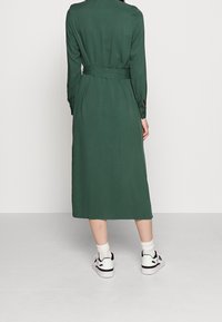 Lång, grön klänning med bälte i midjan, långa ärmar och slitsar i ärmsluten, parat med vita sneakers och vita ankelsockor. Slät textur.