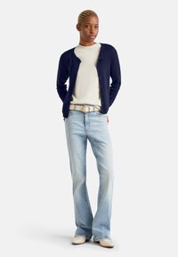 Námořnický kardigán přes krémový top, světle modré rozšířené džíny a béžové boty. Mezi prvky patří knoflíky na kardigánu a kulatý opasek.