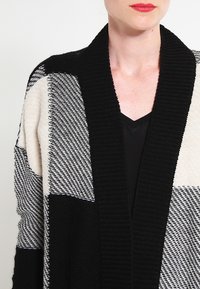 Cardigan nero e crema con motivo a scialle, caratterizzato da un maglione a trama testurizzata. Il design include motivi a blocchi e una vestibilità ampia.