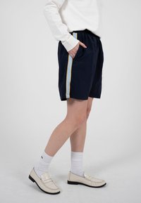 Personne portant un short bleu marine avec des rayures latérales bleu clair et jaune, un haut à manches longues blanc, des chaussettes blanches et des mocassins crème, debout de profil.