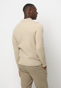 Beige Rippenstrickpullover mit Kragen, taillierter Passform und langen Ärmeln. Getragen mit khakifarbigen Hosen, vor einem schlichten Hintergrund.