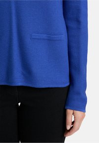 Blauer strukturierter Pullover mit rundem Halsausschnitt, langen Ärmeln und einer Fronttasche. Der Pullover wird mit schwarzen Hosen kombiniert.