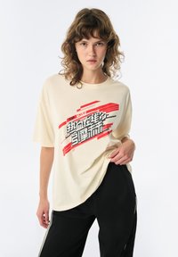 Crème-kleurige oversized t-shirt van zachte stof, met gedurfde rode en zwarte tekst en grafische strepen; gedragen met zwarte broek.
