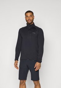 Zwart sporttop met lange mouwen en een ritssluiting aan de kraag, met een gestructureerde stof en contrasterende stikdetails, gecombineerd met donkere shorts.