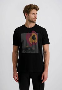 Mann trägt ein schwarzes Pierre Cardin T-Shirt mit abstraktem rot, orange und schwarzem Design und schaut nach rechts vor einem schlichten, hellen Hintergrund.