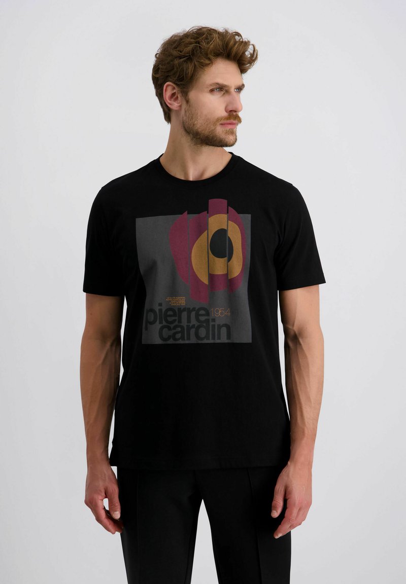 Mann trägt ein schwarzes Pierre Cardin T-Shirt mit abstraktem rot, orange und schwarzem Design und schaut nach rechts vor einem schlichten, hellen Hintergrund.