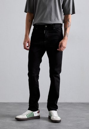 Schwarze Denim-Jeans mit schmaler Passform, mit einem Knopfverschluss und Taschen. Getragen mit einem grauen übergroßen T-Shirt und weißen Sneakers mit grünen Akzenten.