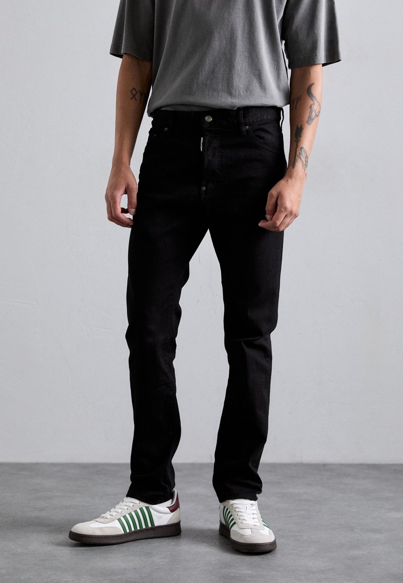 Schwarze Denim-Jeans mit schmaler Passform, mit einem Knopfverschluss und Taschen. Getragen mit einem grauen übergroßen T-Shirt und weißen Sneakers mit grünen Akzenten.