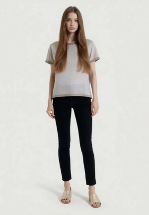 Giovane donna con capelli lunghi e lisci che indossa una maglietta beige a maniche corte, pantaloni neri aderenti e sandali beige con tacco largo e punta aperta, in piedi.