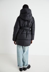 Zwarte gewatteerde jas met een capuchon, een ceintuur in de taille en een gestikt ontwerp. Draagt over lichtblauwe wijde jeans en zwarte schoenen.