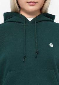 Hoodie vert foncé avec cordon de serrage, poche kangourou et petit logo blanc sur la poitrine. Fabriqué en tissu doux et texturé.