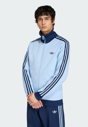 Ung mand med krøllet hår iført en lyseblå Adidas træningsjakke med mørkeblå striber og matchende mørkeblå bukser.