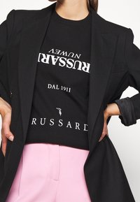 Camiseta negra con texto y logo en blanco, llevada bajo un blazer negro. Combinada con pantalones rosa claro, mostrando un look casual pero estructurado.