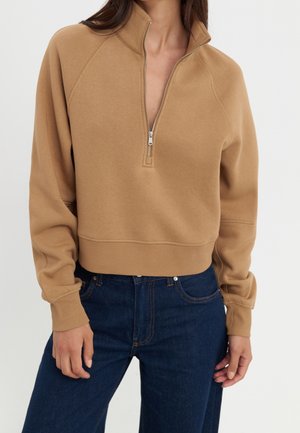 Kobieta ubrana w polar w kolorze wielbłądzim z zamkiem do ćwiartki i ciemnoniebieskie dżinsy, stojąca na tle jednolitego jasnego tła.