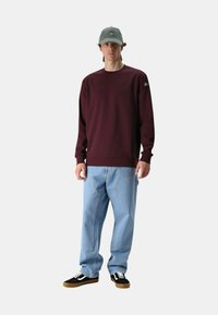 Felpa bordeaux, vestibilità rilassata con orlo a coste, abbinata a jeans baggy azzurri. Sneakers nere con lacci bianchi e cappellino grigio.