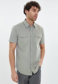 Camisa de manga corta abotonada en tela gris claro, con dos bolsillos en la parte frontal y un dobladillo curvado. Detalle de pequeño logo en el dobladillo.