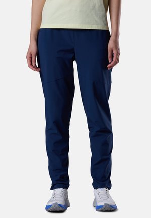 Person iført marineblå bukser, hvide og blå sports sko og en lysegul skjorte, står med hænderne afslappet ned langs siderne.