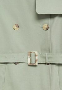 Trench-coat vert clair avec boutons en écaille de tortue et boucle de ceinture assortie, gros plan sur les détails et les coutures à l'avant.
