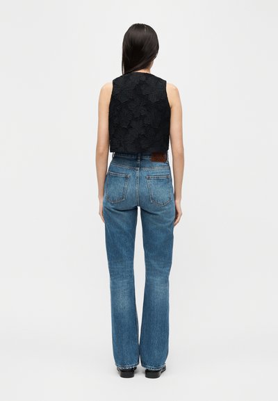 Γυναίκα με μακριά σκουρόχρωμα μαλλιά, που φοράει ένα αμάνικο μαύρο δαντελένιο τοπ και ψηλόμεσα μπλε τζιν, στέκεται γυρισμένη με την πλάτη προς τα εμπρός σε λευκό φόντο.