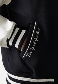 Μαύρο και λευκό μπουφάν bomber με μάλλινο κορμό, δερμάτινα μανίκια, ριγέ ριμπ μανσέτες και κεντημένο το λογότυπο "New York Yankees" στην τσέπη.