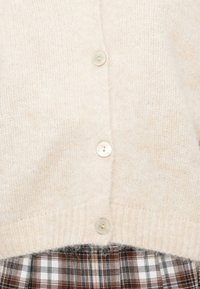 Miękki beżowy sweter z dzianiny z trzema kremowymi guzikami, zestawiony z brązową, białą i czarną spódnicą w kratę.