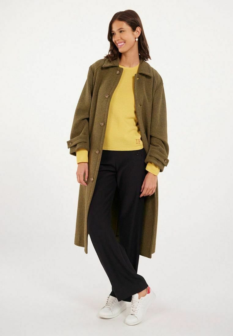 Manteau long olive vert avec des boutons, sur un pull jaune, associé à un pantalon large noir et des baskets blanches. Tissu lisse, coupe décontractée.