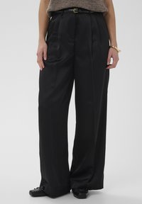 Črne široke hlače iz gladke tkanine, s pregibi in pasom z zlato zaponko. Kombinirane s črnimi ravnimi čevlji.