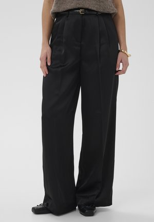 Schwarze Hosen mit weitem Bein aus glattem Stoff, mit Falten und einem Gürtel mit goldener Schnalle. Kombiniert mit schwarzen flachen Schuhen.