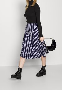 Černý rolák, midi sukně s námořnicko-modrými a bílými pruhy a černé kotníkové boty s šněrováním. V pravé ruce má černou kulatou kabelku.