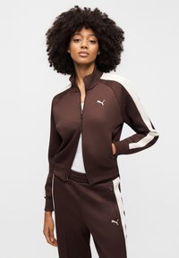 Καφέ ζακέτα φόρμας με φερμουάρ και λευκές ρίγες στα πλάγια, κατασκευασμένη από λείο ύφασμα. Διαθέτει ψηλό γιακά και λογότυπο στο στήθος, σε συνδυασμό με παντελόνι που ταιριάζει.