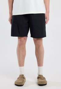 Black cotton shorts with a relaxed fit, rolled hem, and side pockets, paired with beige clogs and white ribbed socks.