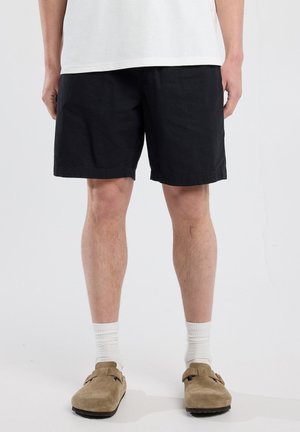 Shorts en coton noir avec une coupe décontractée, ourlet roulé et poches latérales, associés à des sabots beiges et des chaussettes blanches côtelées.