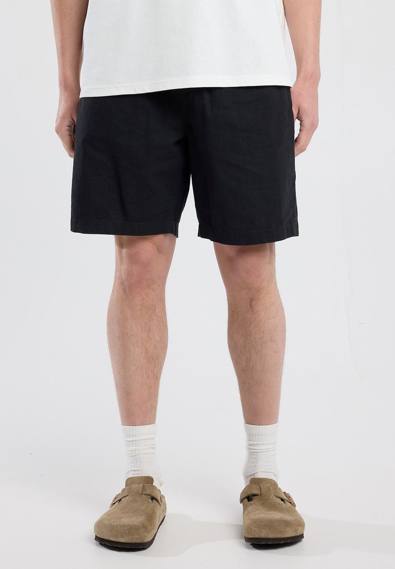 Black cotton shorts with a relaxed fit, rolled hem, and side pockets, paired with beige clogs and white ribbed socks.