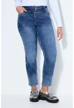 Vrouw met een high-waisted, blauwe spijkerbroek met dubbele knoopsluiting en zilveren plateau loafers, staand tegen een witte achtergrond.