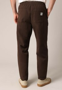 Donkerbruine katoenen broek met een elastische tailleband, twee achterzakken en een subtiele textuur. Schoenen zijn beige met een lichte zool.