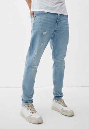 Homme portant un jean bleu clair déchiré et des baskets blanches, debout avec une main dans la poche devant un fond blanc uni.