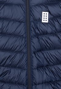 Sötétkék puffos mellény steppelt mintával és vízszintes csíkokkal. Fekete cipzárral és egy kis fehér címkével rendelkezik, hat kerek lyukkal.