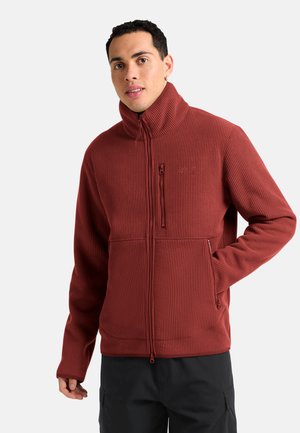 Junger Mann trägt eine rostfarbene, gerippte Reißverschlussjacke mit hohem Kragen und schwarze Hose, steht vor einem schlichten Hintergrund.