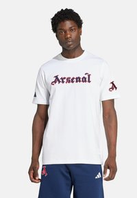 T-shirt blanc à manches courtes avec "Arsenal" en lettres gothiques rouges sur la poitrine et un "A" rouge sur la manche gauche. Matière en coton.